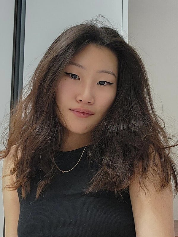 A young woman with long dark hair smiling at the camera, wearing a simple black tank top against a plain indoor background.