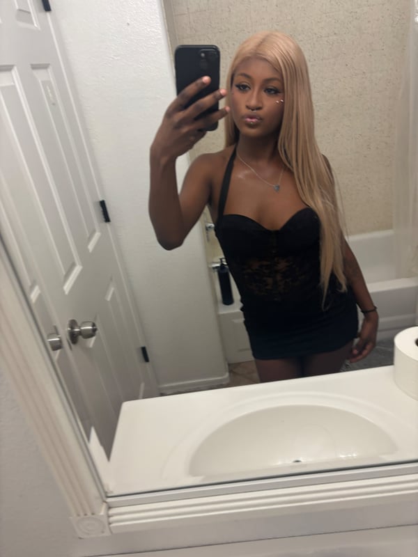 A young woman taking a mirror selfie in a bathroom, wearing a black lace top and short skirt.