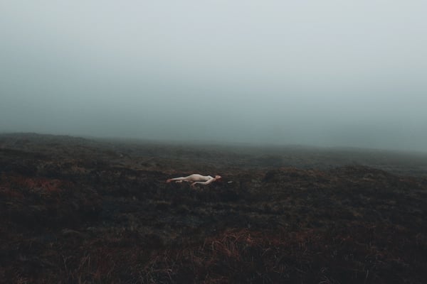 A person lying on a misty grassy hillside in a foggy natural landscape.