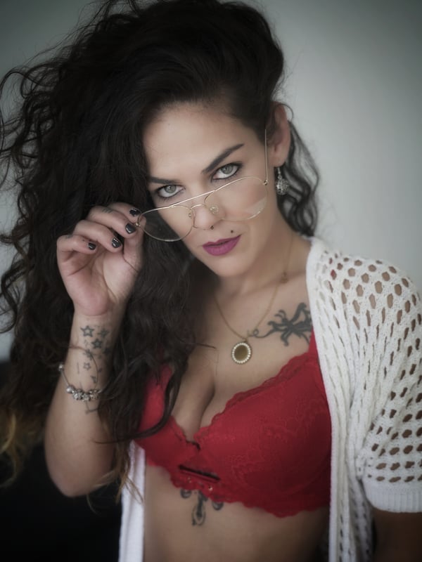 A woman with wavy dark hair and tattoos posing confidently while holding glasses, wearing a red lace top under a white open cardigan.