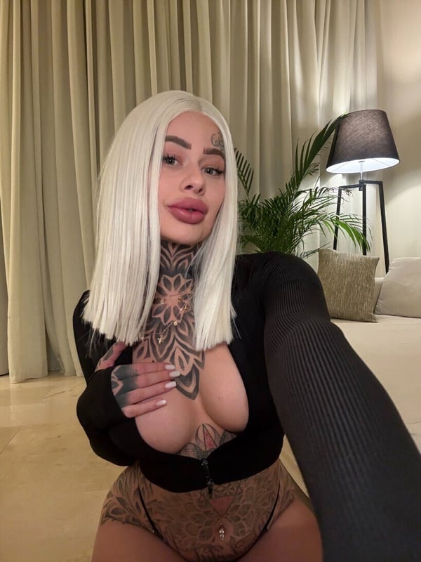 A woman with platinum blonde hair and tattoos stands in a stylish room with curtains, plants, and warm lighting, wearing a sheer top.