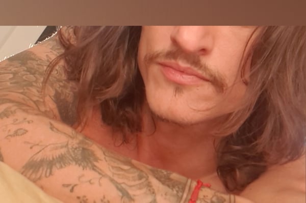 Close-up portrait of a man with long hair, mustache, and extensive tattoos on his shoulders and arms, smiling softly.