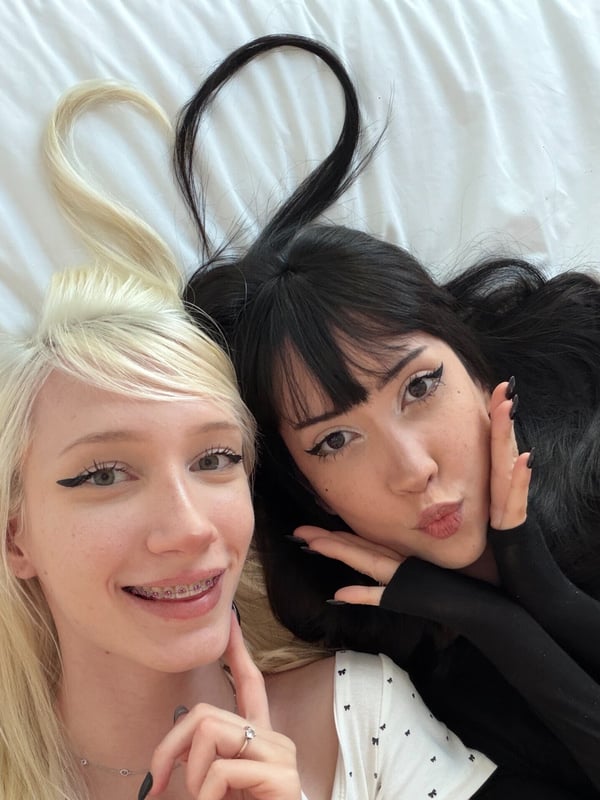 Two young women lying on a bed, smiling and making playful faces with long blonde and black hair.