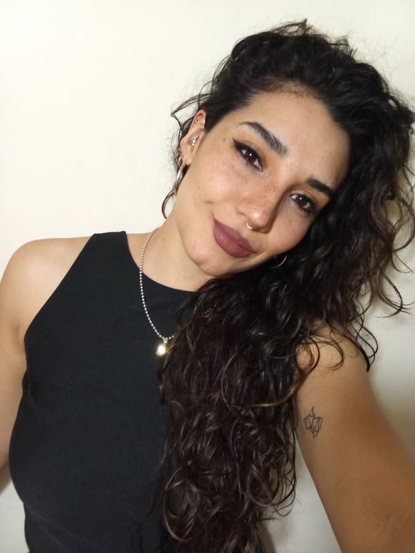 Young woman with long wavy dark hair, wearing a black sleeveless top and gold necklace, posing with a smile against a plain background.