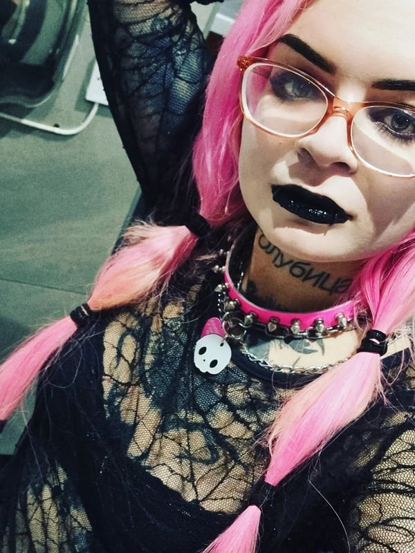 A woman with pink hair in pigtails, wearing glasses, black lipstick, a choker necklace, and sheer black clothing, posing confidently indoors.
