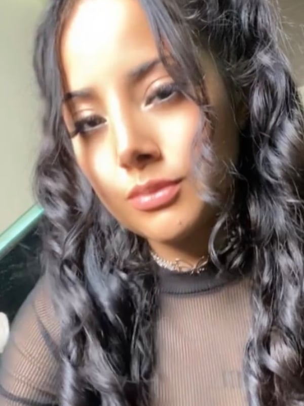 Young woman with long dark curly hair in pigtails, wearing a sheer black top, posing closely with a neutral expression.
