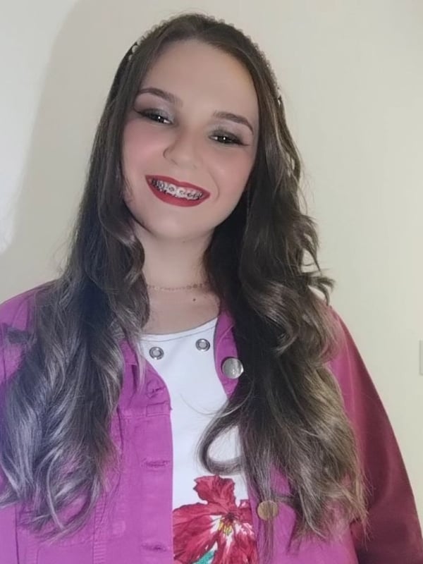 Young woman with braces smiling at the camera, wearing a pink jacket over a white top with wavy brown hair.