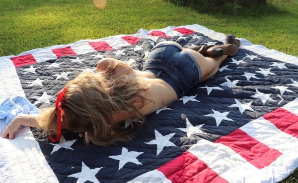 A woman lying on a patriotic quilt on the grass, wearing denim shorts and cowboy boots, with blonde hair and a red headband.