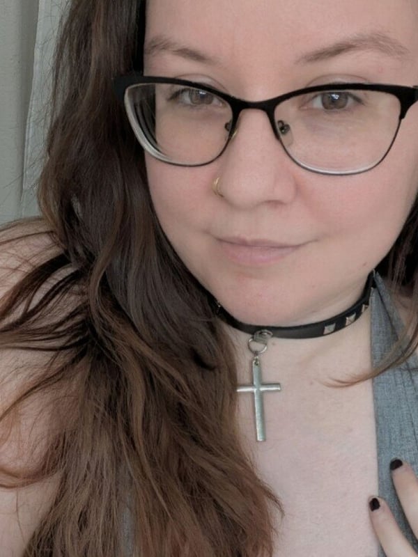 Close-up portrait of a woman with glasses, long brown hair, a choker necklace, and a cross pendant, smiling softly.