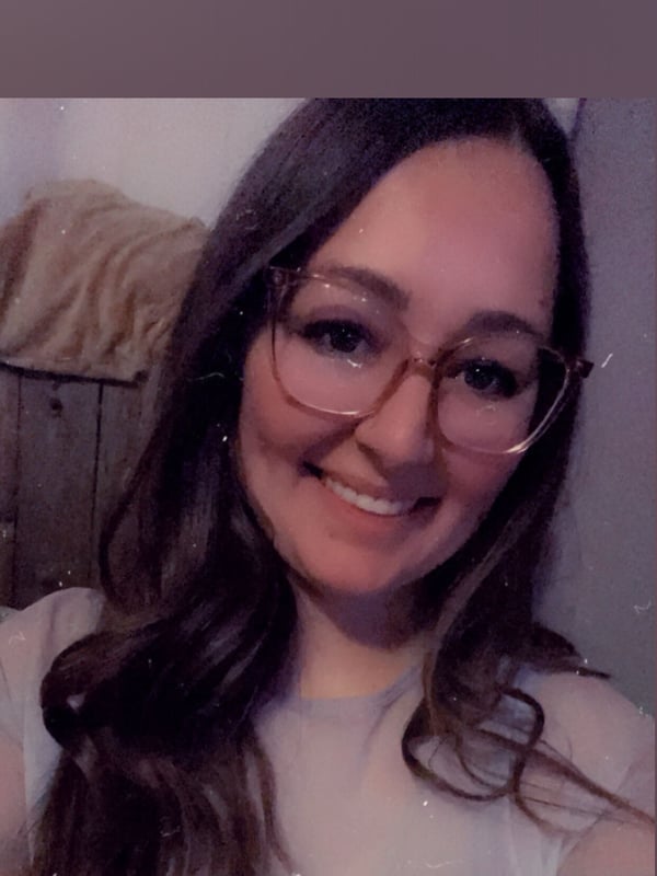 A smiling woman with long wavy hair and glasses, posing casually in a light-colored top.