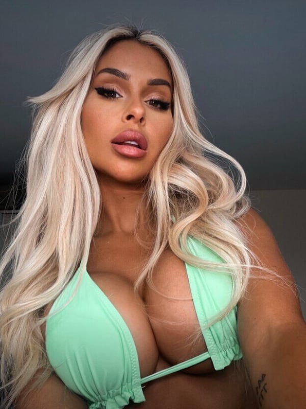 A close-up selfie of a woman with long blonde hair and bold makeup wearing a green bikini top.
