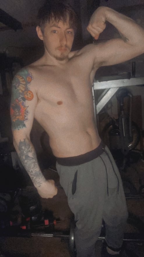 A young man with tattoos flexing his muscles shirtless in a gym setting, wearing gray sweatpants.