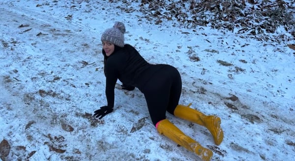 A woman in black leggings and a gray beanie poses on her hands and knees in a snowy outdoor setting, wearing yellow boots.
