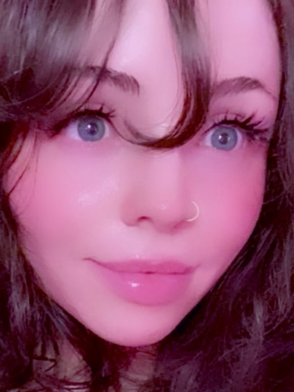 Close-up portrait of a young woman with long brunette hair, blue eyes, and a nose piercing, smiling softly with pink blush tones in a filtered photo.