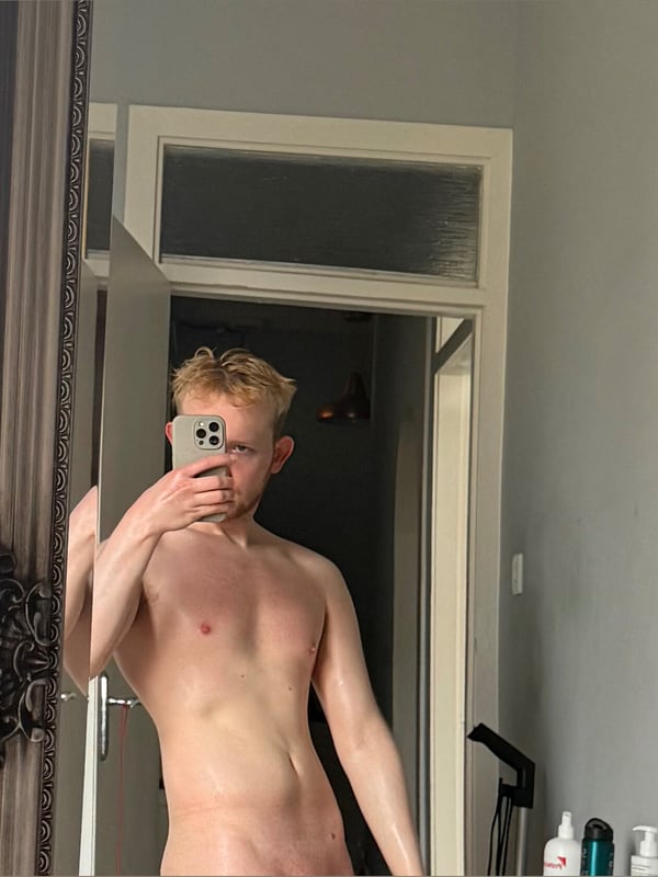 A young man with blonde hair taking a mirror selfie in a bathroom, standing shirtless near a door.