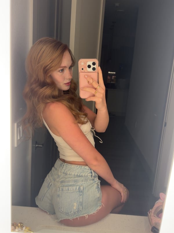 Young woman with blonde hair taking a selfie in a black halter top inside a bedroom with warm lighting.