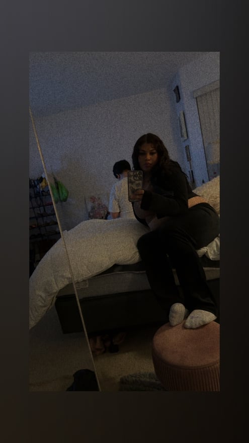 Young woman sitting on a bed in a bedroom, wearing casual black pants and white socks, with another person visible in the background mirror selfie.