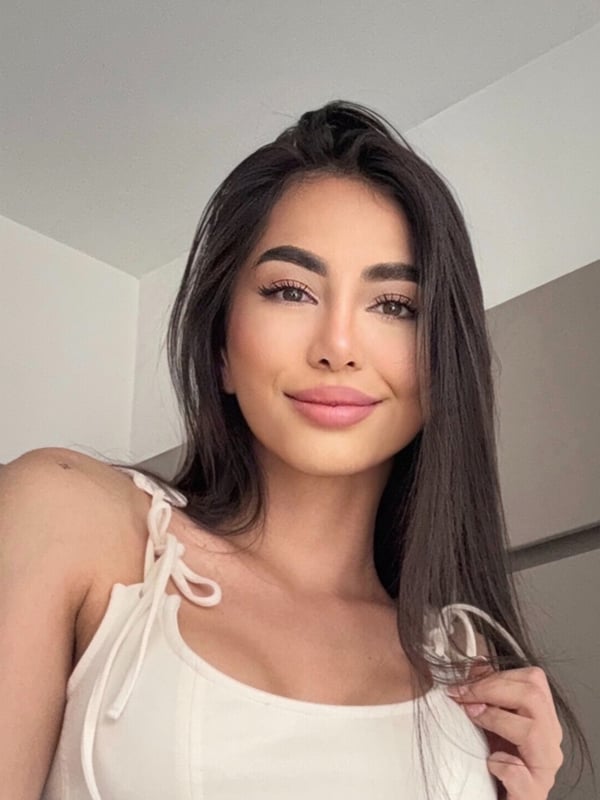 A young woman with long dark hair smiling at the camera, wearing a white top with thin straps in a softly lit indoor setting.