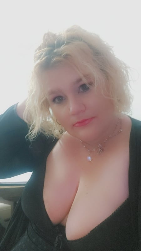 A woman with blonde curly hair smiling confidently while wearing a black top with a necklace, posing indoors.