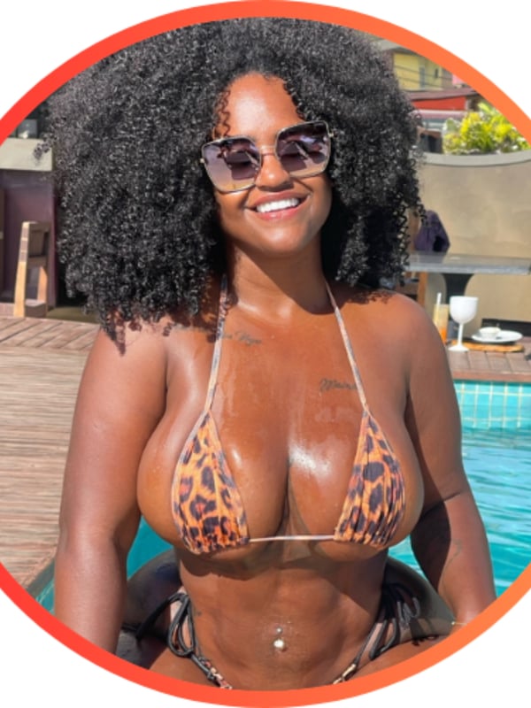 A smiling woman with curly hair wearing sunglasses and a leopard print bikini sits poolside.