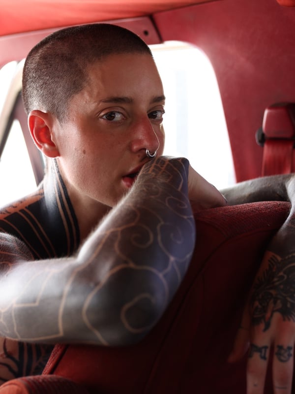 Person with short hair and tattoos posing thoughtfully inside a red car interior.