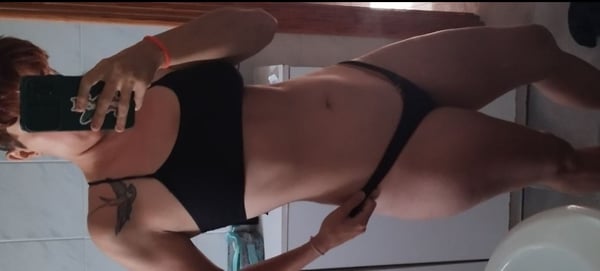 Woman lying on her side in a bathroom mirror selfie, wearing a black bikini top and bottoms, holding her phone.