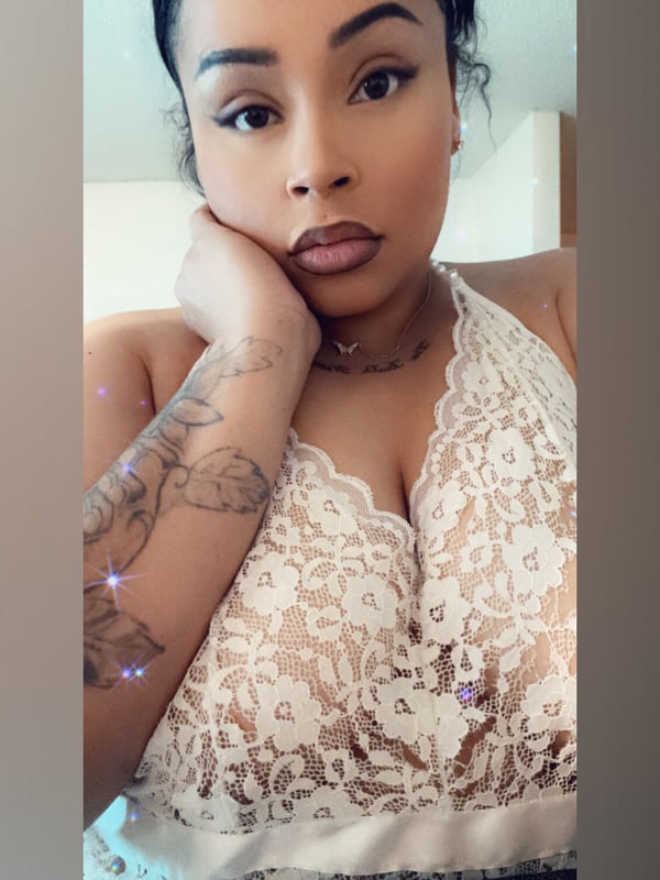 A woman with dark hair and tattoos posing in a white lace top, hand on her chin, against a light indoor background.