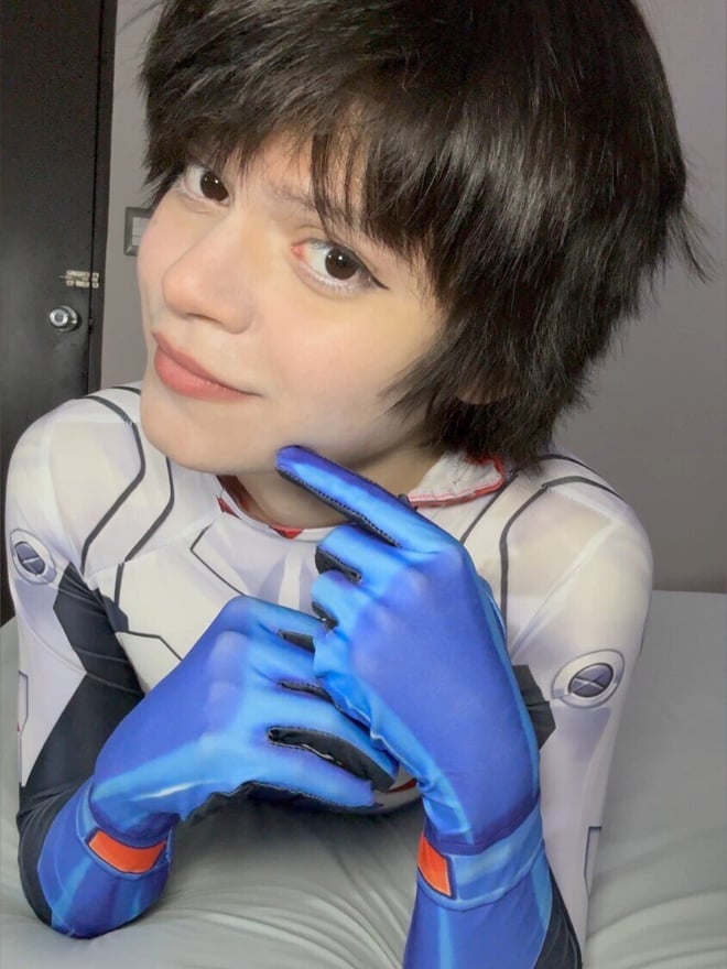 Person with short black hair wearing a white and blue bodysuit with blue gloves, smiling at the camera indoors.