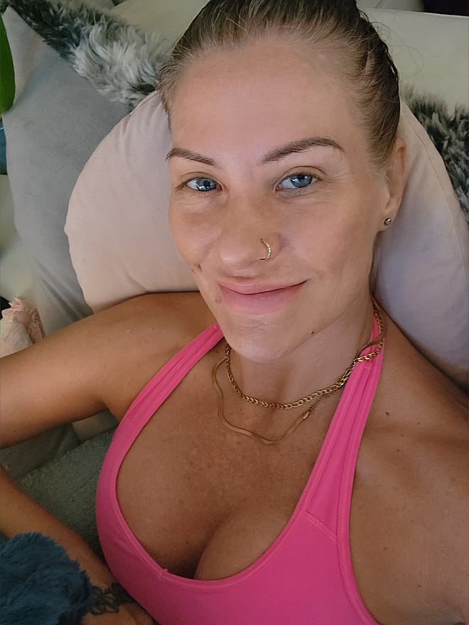A woman with light brown hair smiling on a bed, wearing a pink tank top and layered necklaces in a relaxed indoor environment.