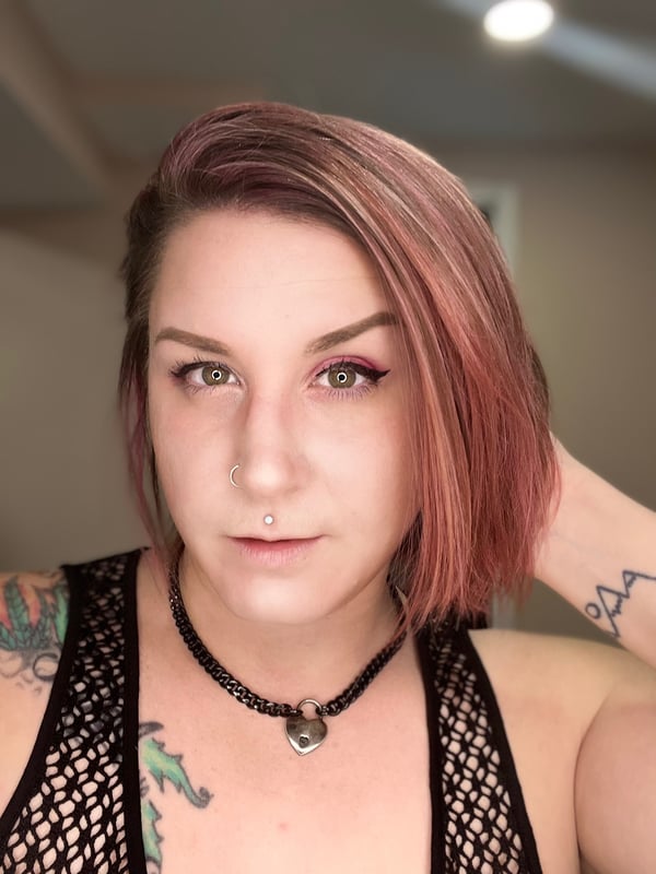 Close-up portrait of a woman with short pinkish hair, green eyes, facial piercing, and tattoos, wearing a black choker and mesh top.