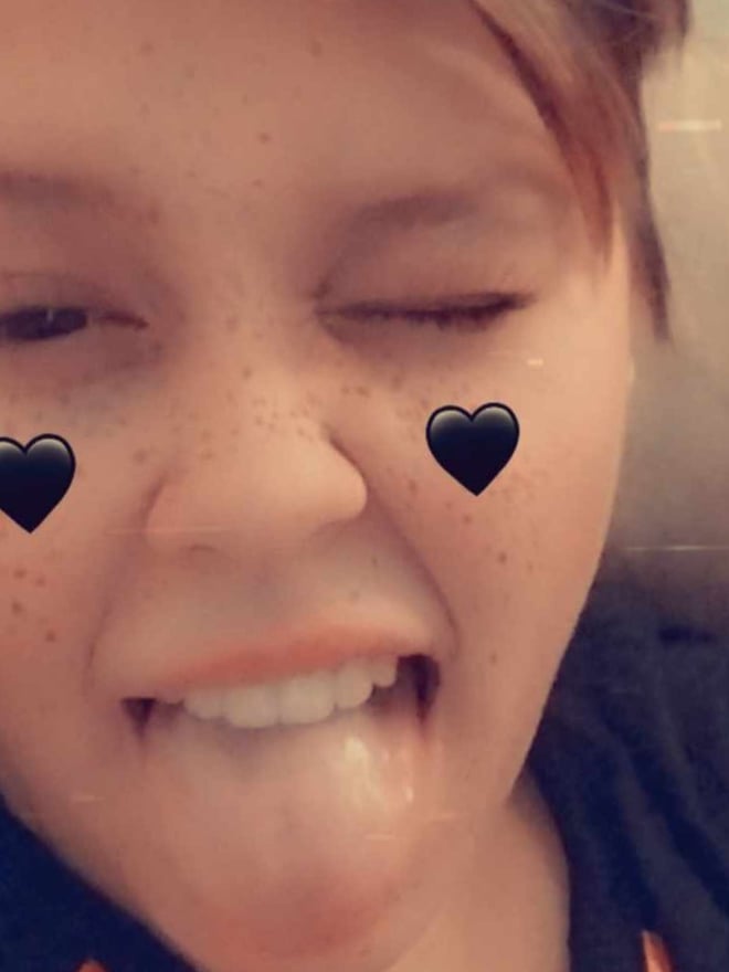 A young person with short brown hair making a playful face with tongue out and heart emojis over eyes, wearing an orange top.