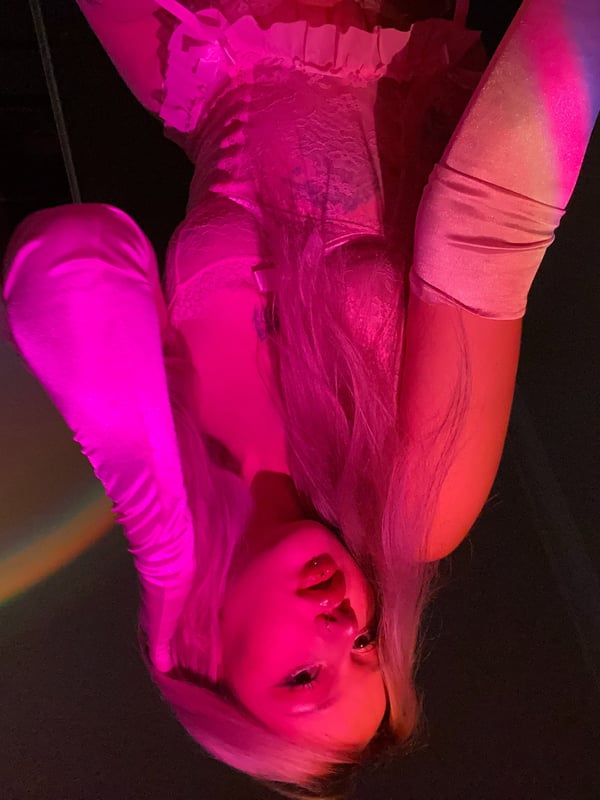 A young woman with long blonde hair in lingerie posing playfully upside down under colorful lighting.
