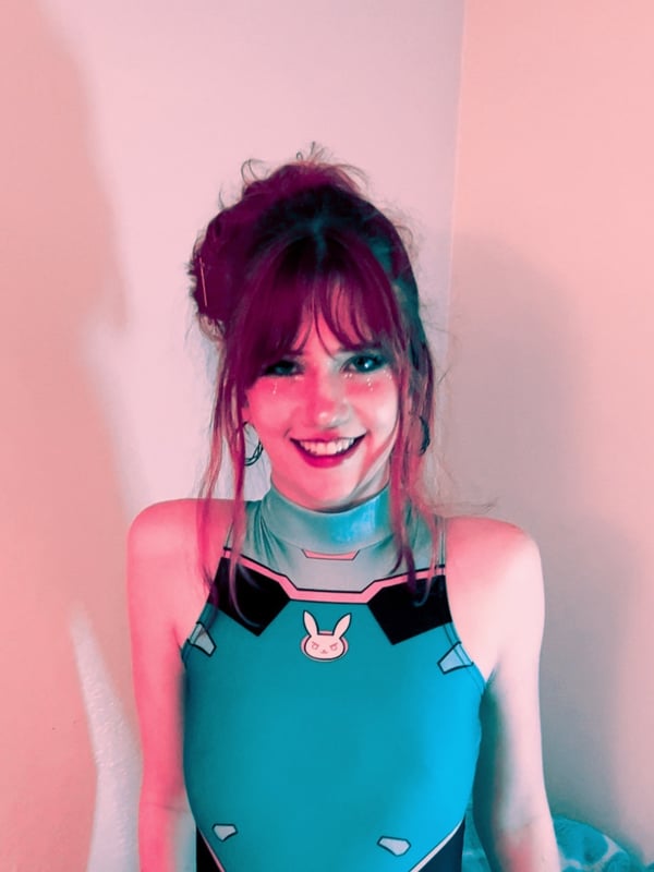 Young woman with reddish-brown hair dressed in a blue cosplay outfit with bunny ears, smiling in a room with pink lighting.