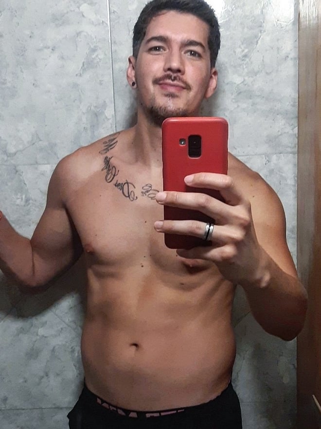 A man taking a shirtless selfie in a bathroom, holding a phone and showing tattoos on his chest.