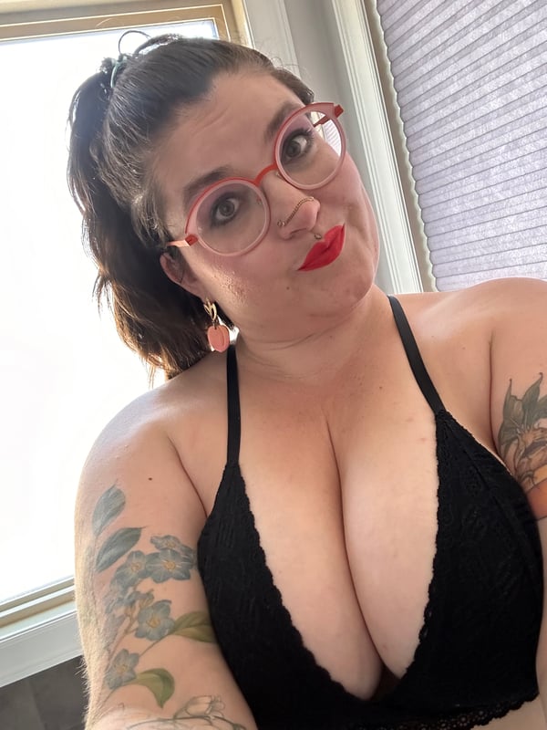 A woman with dark hair in a ponytail and red glasses, smiling at the camera while wearing a black lace top near a window.