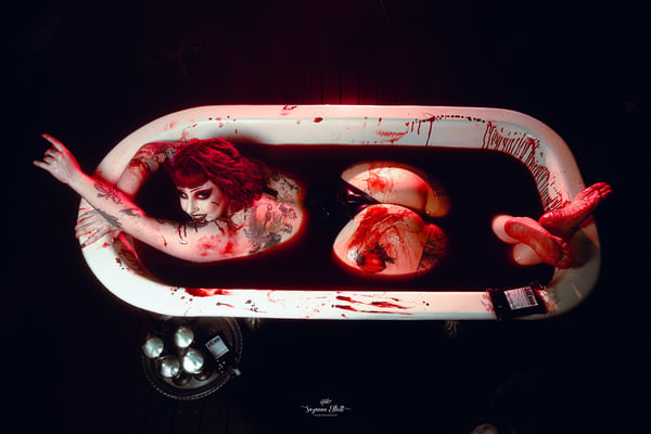 A performer in dramatic red makeup and hair poses artistically in a bathtub with theatrical red lighting and props on a dark background.