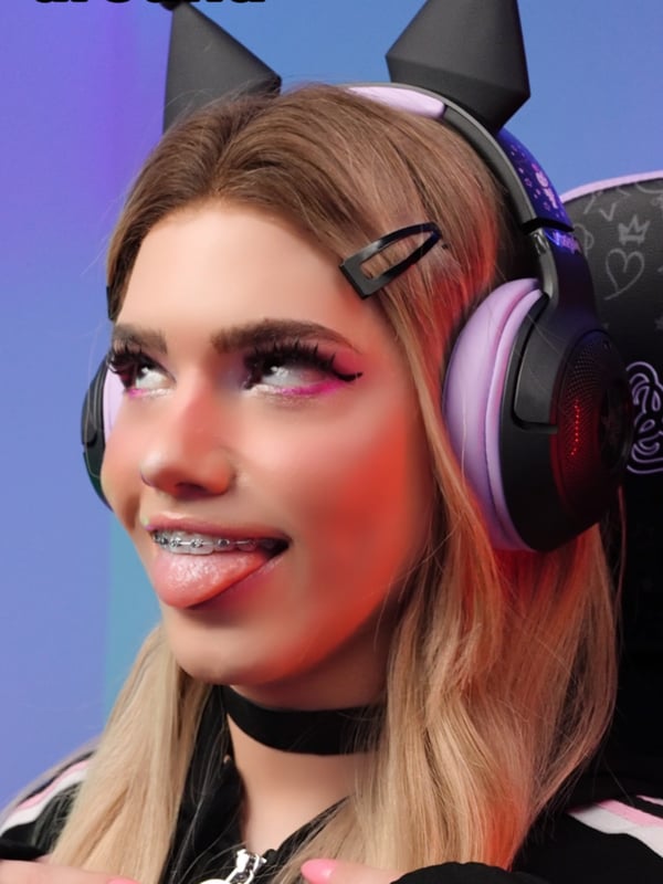 Person with long blonde hair, cat ear headphones, and gaming headset sitting in a gaming chair, wearing a choker and gloves while making a playful expression.