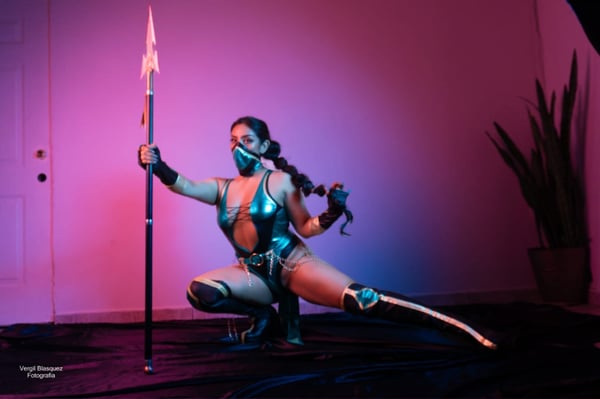 A woman in a shiny green costume and mask poses dynamically with a spear in a room with pink lighting and plants.