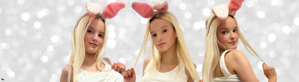 Three young women with blonde hair wearing white tops and pink bunny ears, posing playfully against a sparkly gray background.
