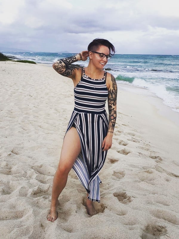 A smiling woman with short hair and tattoos stands on a sandy beach in a striped dress, with ocean waves in the background.