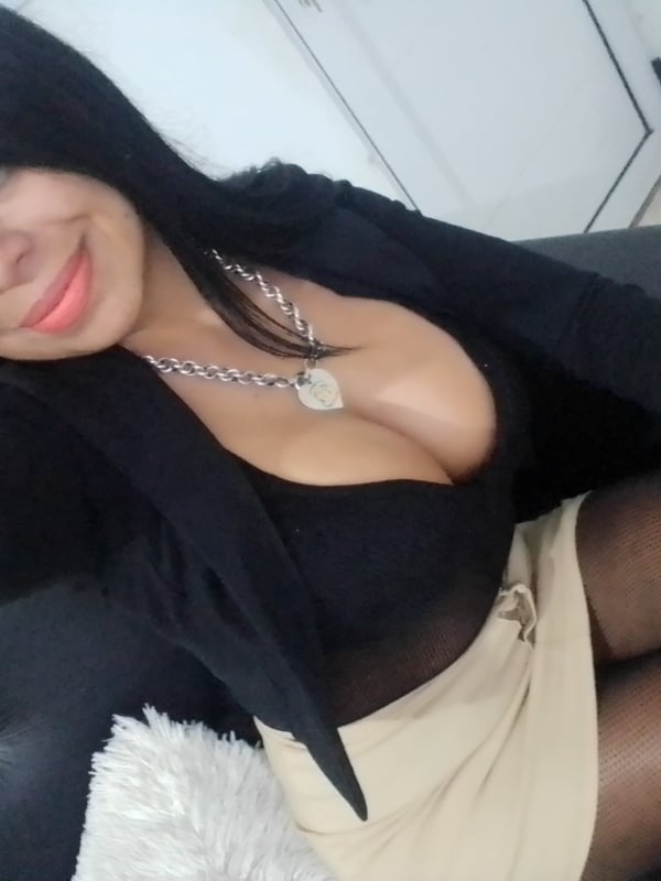 Woman with long dark hair smiling at the camera in a black blazer, low-cut top, beige skirt, and sheer stockings while seated on dark furniture.