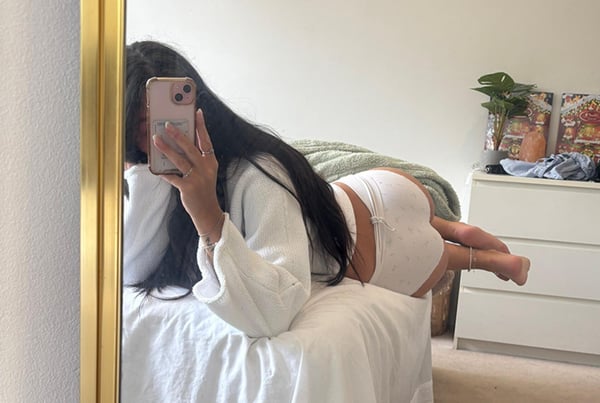 Young woman lying on a bed in a cozy bedroom, taking a mirror selfie while wearing a white robe and shorts.