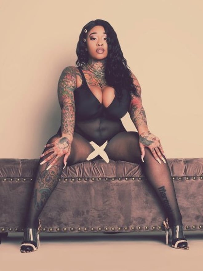 A woman with braided hair and tattoos sits on a bench in a black bodysuit and stockings, posing confidently.