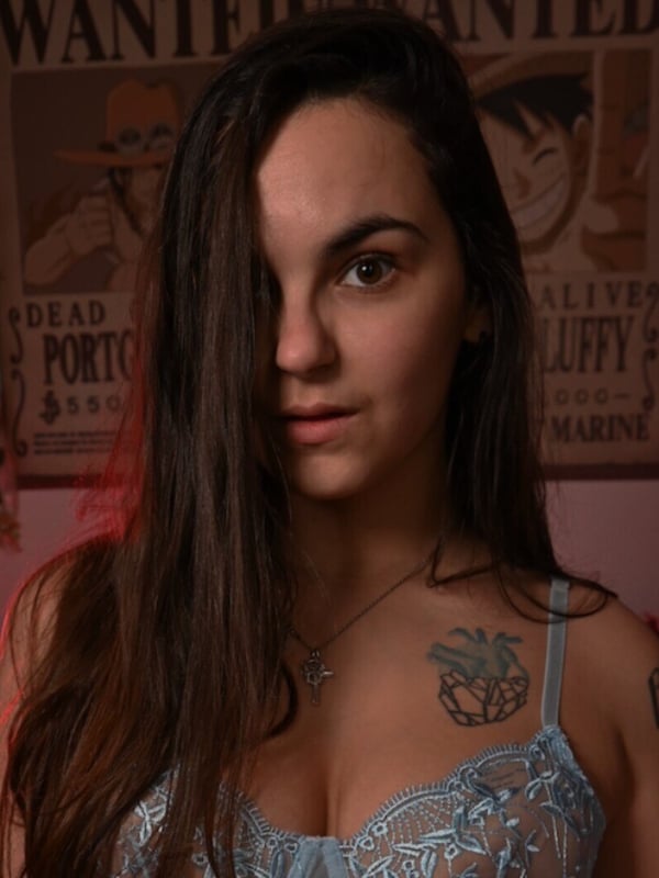 A woman with long dark hair standing in front of a vintage movie poster, wearing lace lingerie in a dimly lit room.