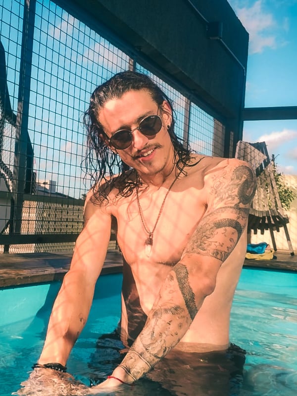 Man with long wet hair and sunglasses sitting by a pool in swim trunks, with tattoos visible on his arms, against a metal fence and blue sky background.