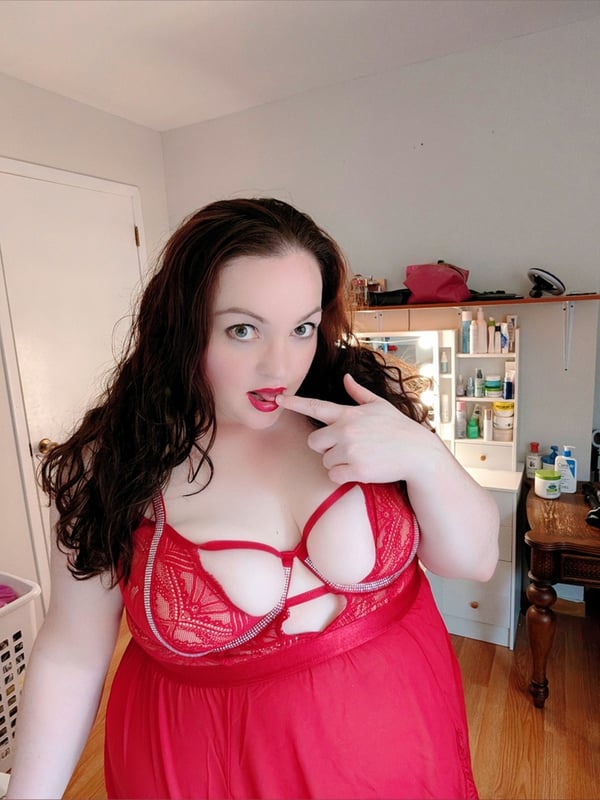 A woman with long wavy dark hair standing in a room, wearing a red lace lingerie outfit, making a playful gesture with her finger.