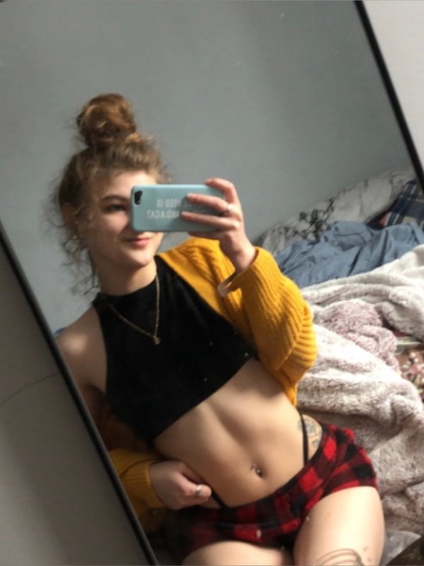 A young woman taking a mirror selfie, smiling with a phone in hand, dressed in a black crop top, orange cardigan, and plaid shorts.