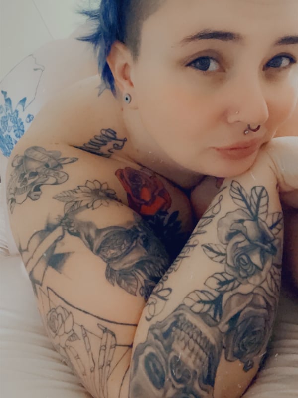 A woman with blue spiked hair and extensive tattoos poses sideways on a white surface, wearing earrings.