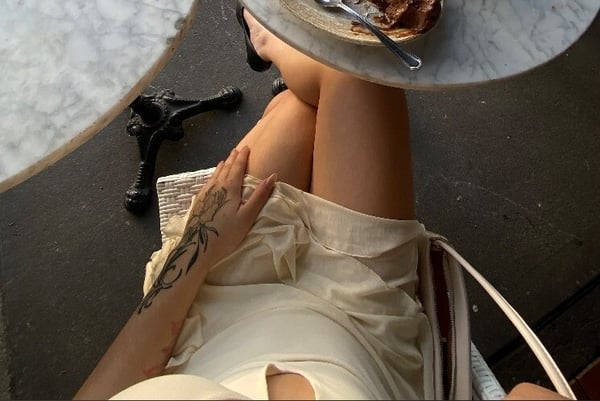 A woman sitting at an outdoor cafe table with dessert, wearing a light dress and showing tattoos on her arm.