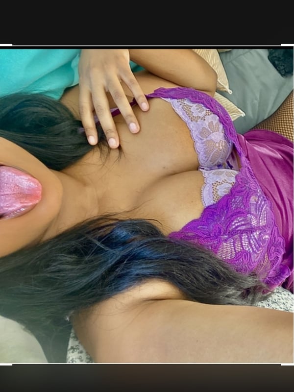 Close-up selfie of a woman in purple lingerie making a playful expression on a bed.